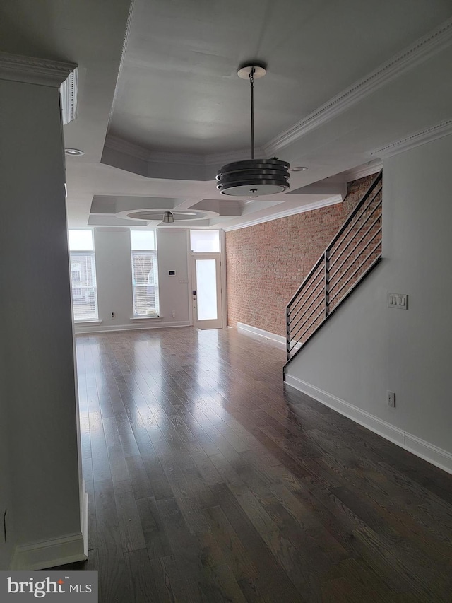 unfurnished living room featuring brick wall, dark wood-style flooring, stairs, ornamental molding, and a tray ceiling