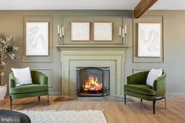 living area featuring a brick fireplace, beamed ceiling, baseboards, and wood finished floors