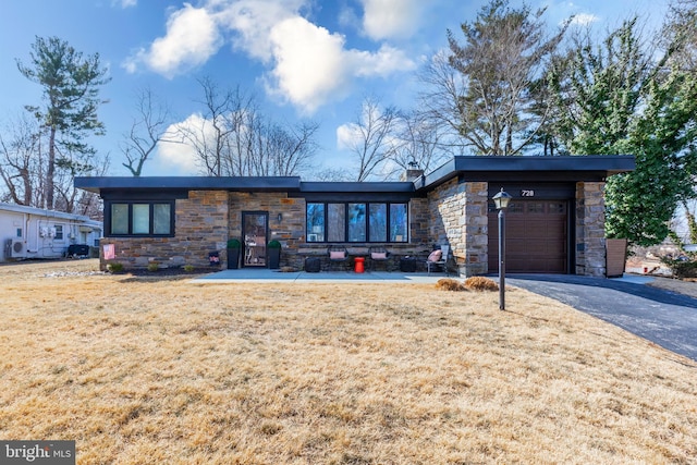 mid-century home featuring a chimney, an attached garage, stone siding, driveway, and a front lawn