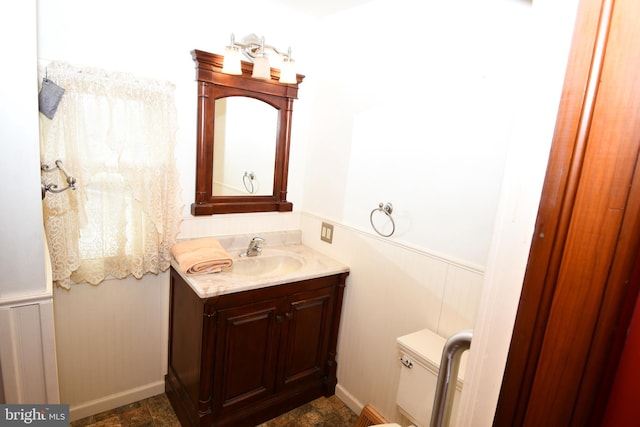 bathroom with a wainscoted wall, vanity, stone finish floors, and toilet