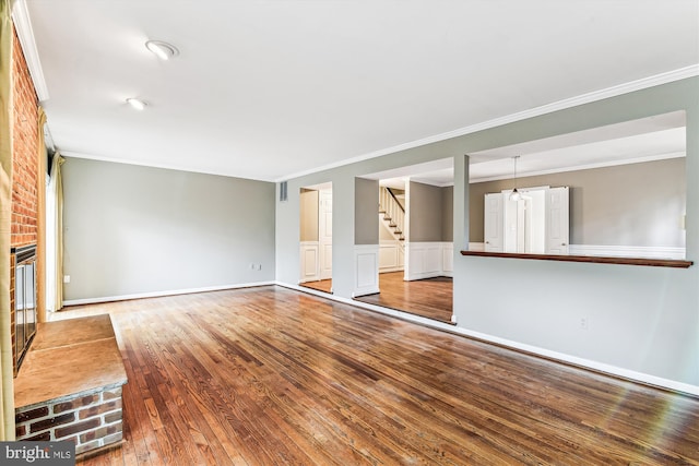 unfurnished living room with crown molding, wood-type flooring, a brick fireplace, wainscoting, and stairs