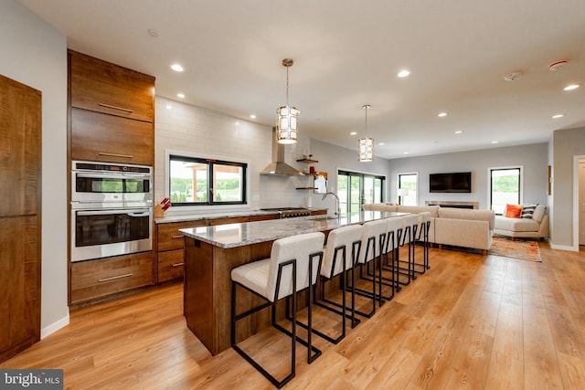 kitchen with brown cabinetry, appliances with stainless steel finishes, a breakfast bar area, light wood-type flooring, and wall chimney range hood