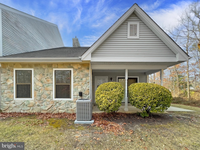 rear view of property with central AC unit, stone siding, a chimney, roof with shingles, and a porch