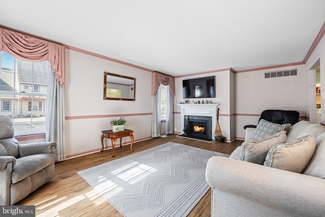 living area featuring a warm lit fireplace, wood finished floors, visible vents, baseboards, and ornamental molding