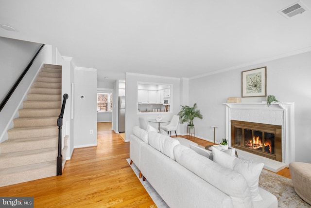 living room with visible vents, a brick fireplace, stairway, ornamental molding, and light wood-style flooring