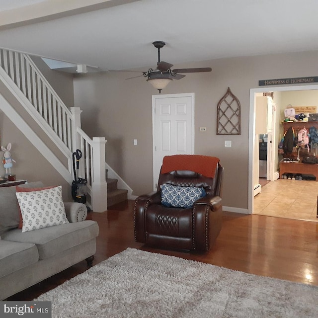 living area with ceiling fan, a baseboard heating unit, wood finished floors, baseboards, and stairway