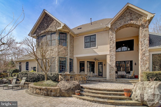 rear view of house with roof with shingles, stucco siding, a ceiling fan, a patio area, and stone siding