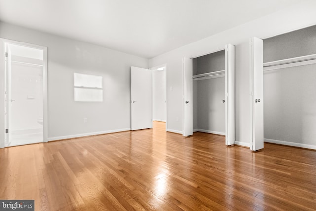 unfurnished bedroom with wood finished floors, two closets, connected bathroom, and baseboards