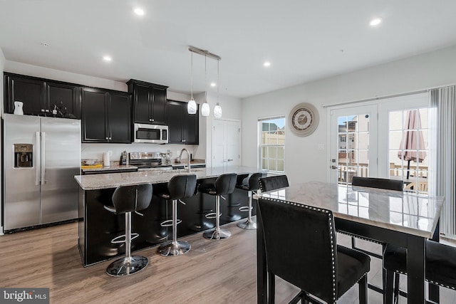 kitchen with a breakfast bar area, light wood finished floors, stainless steel appliances, a kitchen island with sink, and dark cabinets