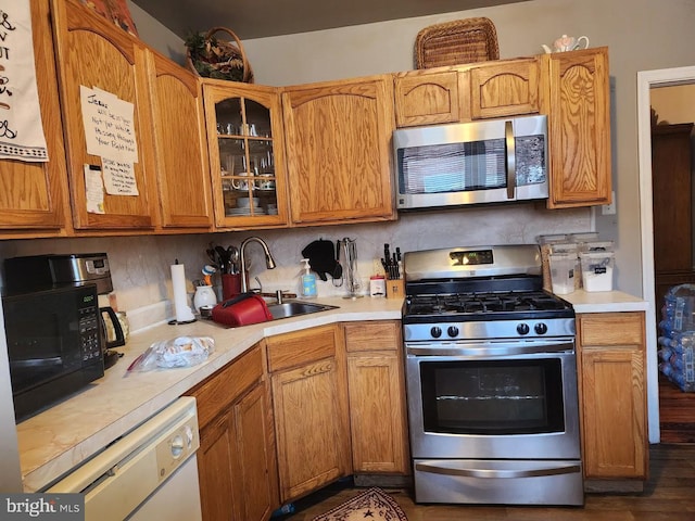 kitchen with stainless steel appliances, a sink, light countertops, dark wood finished floors, and glass insert cabinets