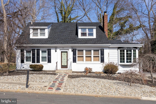 cape cod house with stucco siding, a chimney, and roof with shingles