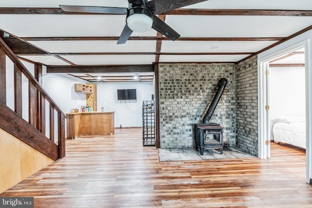 unfurnished living room featuring beamed ceiling, wood finished floors, a wood stove, and a ceiling fan