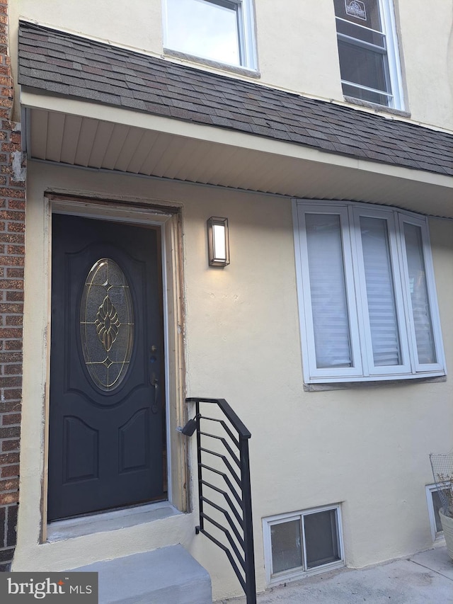 doorway to property featuring brick siding, roof with shingles, and stucco siding