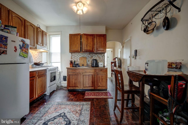 kitchen with white appliances, tasteful backsplash, arched walkways, brown cabinetry, and under cabinet range hood