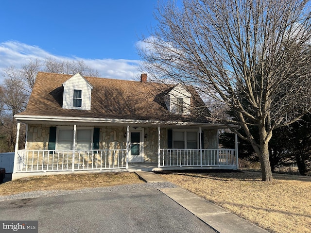 cape cod home featuring stone siding, covered porch, a chimney, and roof with shingles