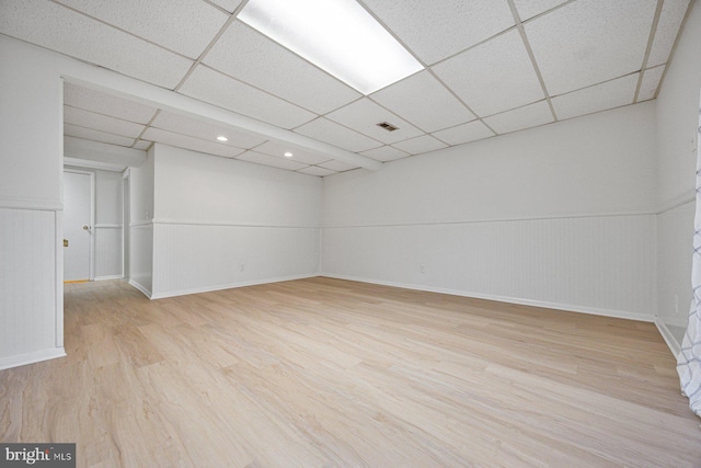 finished basement with wainscoting, light wood-type flooring, and a paneled ceiling