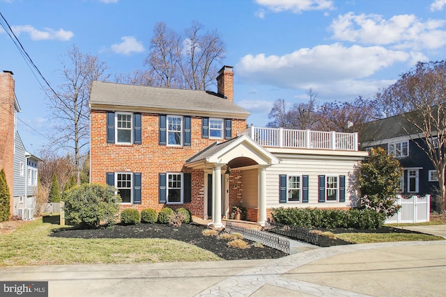 view of front facade featuring a balcony, brick siding, fence, a chimney, and a front yard