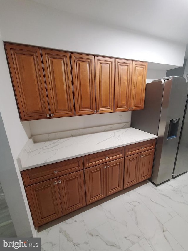 kitchen with brown cabinets, marble finish floor, stainless steel refrigerator with ice dispenser, and light stone countertops