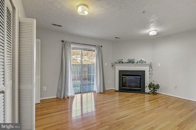 unfurnished living room with a textured ceiling, a fireplace with flush hearth, visible vents, baseboards, and light wood-style floors