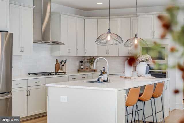 kitchen with stainless steel appliances, tasteful backsplash, light wood-style flooring, a kitchen island with sink, and wall chimney exhaust hood