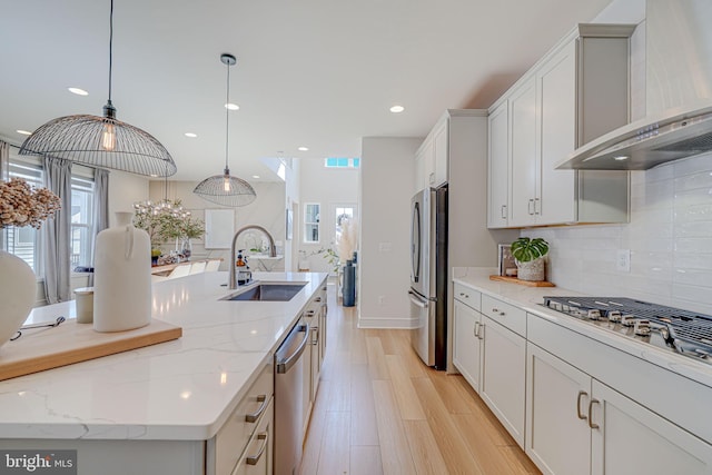 kitchen with decorative backsplash, appliances with stainless steel finishes, a kitchen island with sink, a sink, and wall chimney exhaust hood