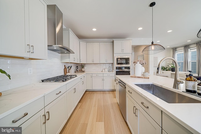 kitchen featuring wall chimney exhaust hood, appliances with stainless steel finishes, hanging light fixtures, light wood-type flooring, and a sink