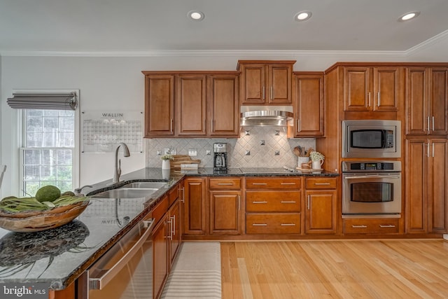 kitchen with brown cabinets, appliances with stainless steel finishes, a sink, dark stone counters, and under cabinet range hood