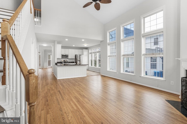 unfurnished living room featuring light wood-style flooring, a fireplace with flush hearth, ceiling fan, high vaulted ceiling, and stairs