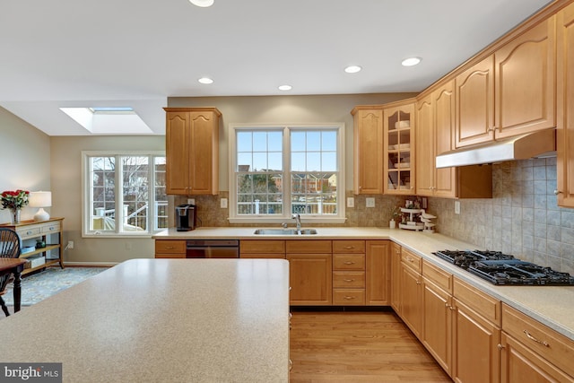 kitchen with light wood-type flooring, black gas stovetop, under cabinet range hood, a sink, and a skylight