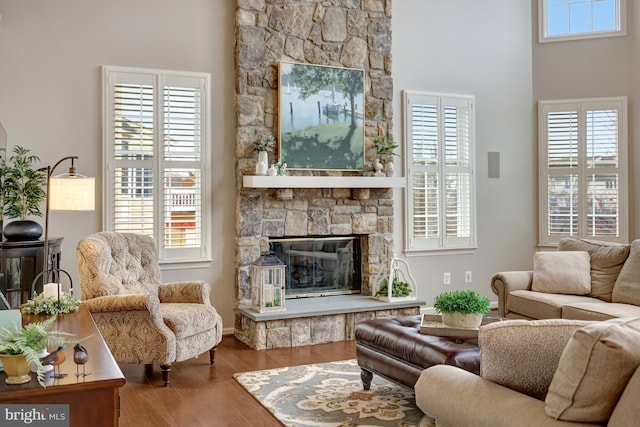 living area with a wealth of natural light, a stone fireplace, wood finished floors, and a towering ceiling