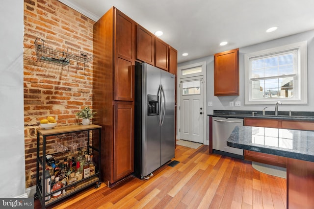 kitchen with dark stone counters, brick wall, appliances with stainless steel finishes, light wood-type flooring, and a sink