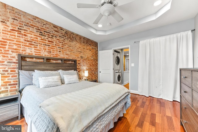 bedroom with ceiling fan, brick wall, wood finished floors, a tray ceiling, and stacked washer and clothes dryer