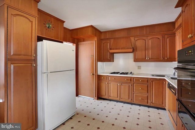 kitchen with gas stovetop, custom exhaust hood, freestanding refrigerator, light floors, and brown cabinetry