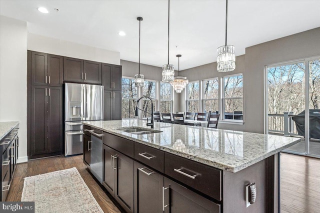 kitchen with an island with sink, a sink, stainless steel appliances, dark brown cabinetry, and dark wood-style flooring