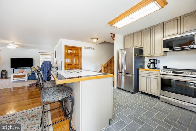 kitchen featuring visible vents, ceiling fan, tile countertops, appliances with stainless steel finishes, and a kitchen breakfast bar