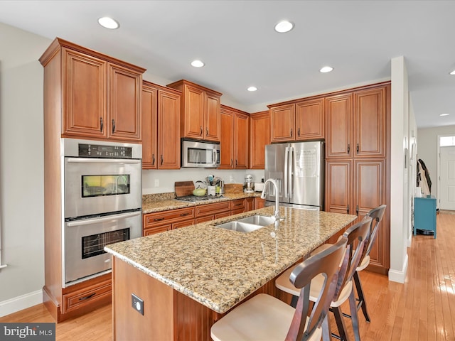 kitchen with a sink, appliances with stainless steel finishes, light stone countertops, light wood finished floors, and brown cabinetry