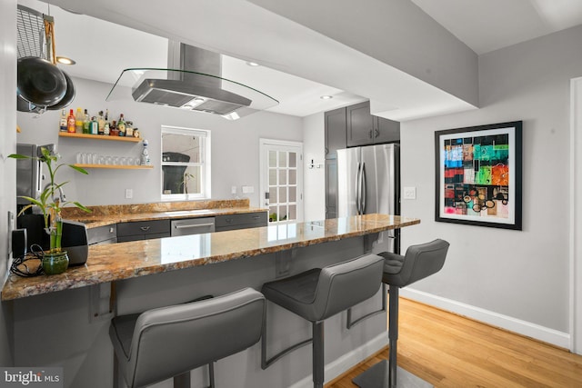 kitchen featuring a breakfast bar area, light stone counters, a peninsula, exhaust hood, and stainless steel appliances