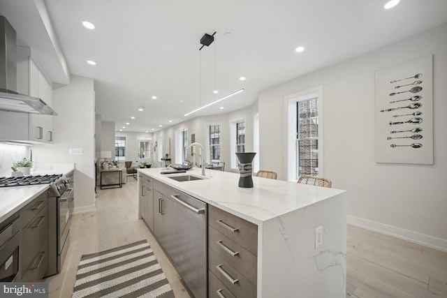 kitchen featuring a center island with sink, appliances with stainless steel finishes, open floor plan, a sink, and wall chimney range hood