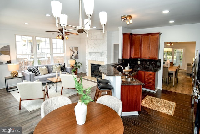 kitchen featuring dark wood finished floors, backsplash, ceiling fan, a stone fireplace, and a peninsula