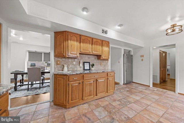 kitchen with visible vents, backsplash, freestanding refrigerator, stone finish flooring, and brown cabinetry