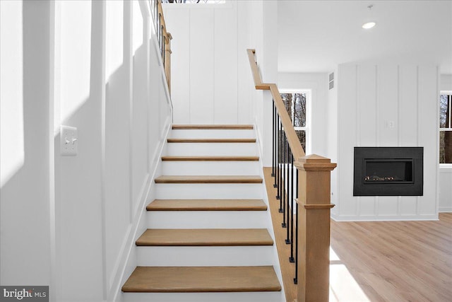 staircase with recessed lighting, a decorative wall, wood finished floors, and a glass covered fireplace