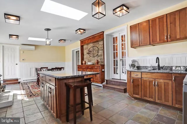 kitchen with a wall unit AC, a wainscoted wall, dark stone counters, decorative light fixtures, and brown cabinets