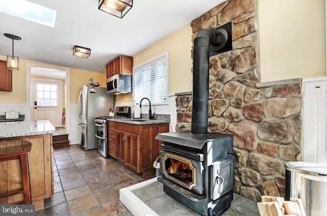 kitchen with a sink, stainless steel appliances, wainscoting, light stone countertops, and a wood stove