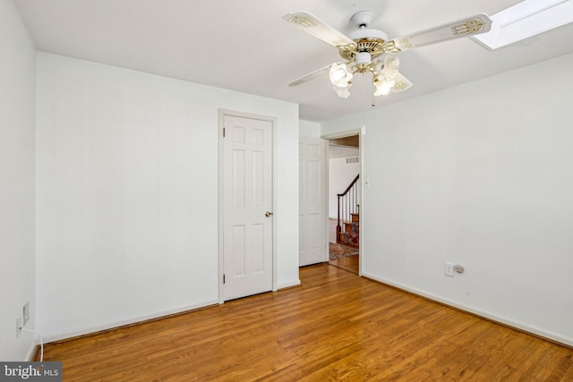 empty room featuring a skylight, a ceiling fan, wood finished floors, baseboards, and stairs