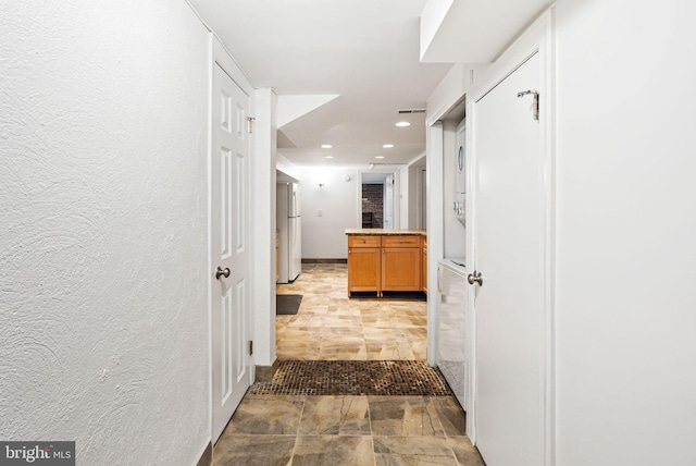 hallway featuring a textured wall, stone finish flooring, stacked washer and clothes dryer, and recessed lighting