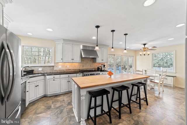 kitchen with wall chimney range hood, butcher block countertops, decorative backsplash, stainless steel appliances, and a sink