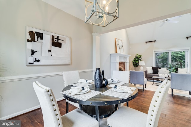 dining space featuring wood-type flooring, ornate columns, vaulted ceiling, a brick fireplace, and ceiling fan with notable chandelier