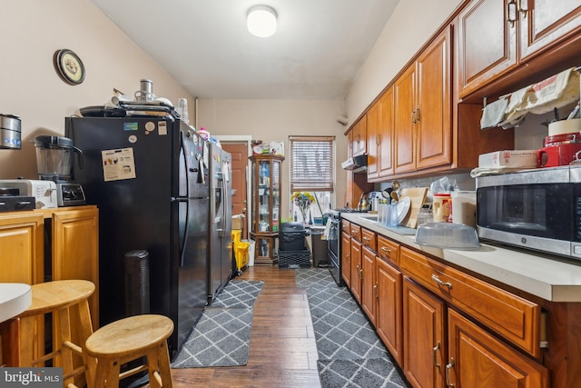 kitchen with brown cabinetry, dark wood-type flooring, stainless steel appliances, light countertops, and under cabinet range hood