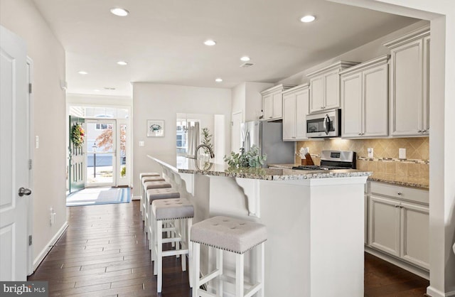 kitchen with a breakfast bar, appliances with stainless steel finishes, backsplash, an island with sink, and dark wood finished floors