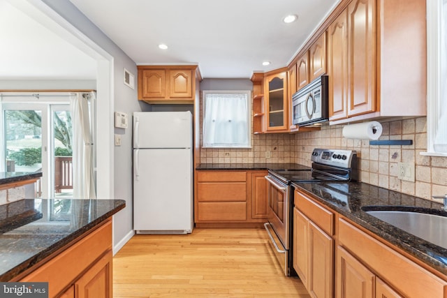 kitchen featuring light wood-style flooring, stainless steel appliances, decorative backsplash, dark stone countertops, and glass insert cabinets